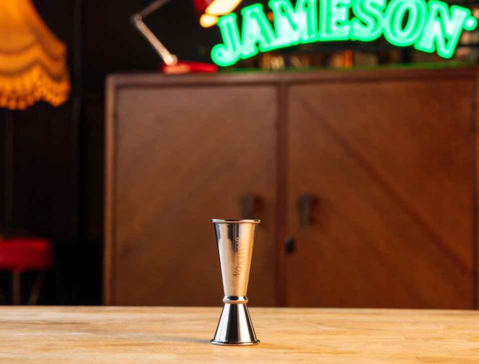 A Jameson branded jigger in front of a Jameson green illuminated sign