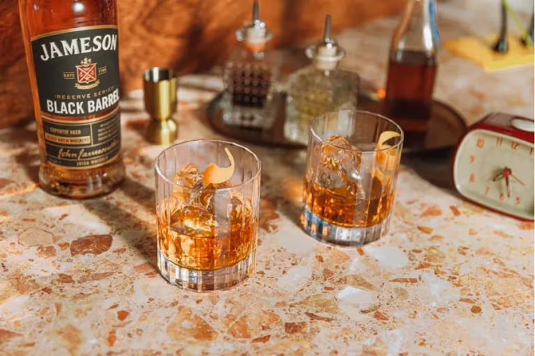 two glasses of whiskey with ice and orange peels on a marble countertop next to a bottle of jameson black barrel and various cocktail making tools